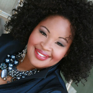 American soprano Karen Slack smiles warmly, with lush curly dark hair, a statement necklace of large gray baubles, and black clothes