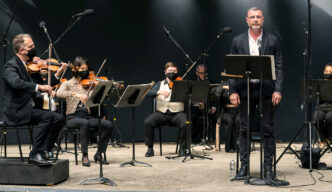 Liev Schreiber and members of Orpheus perform on stage against a black background