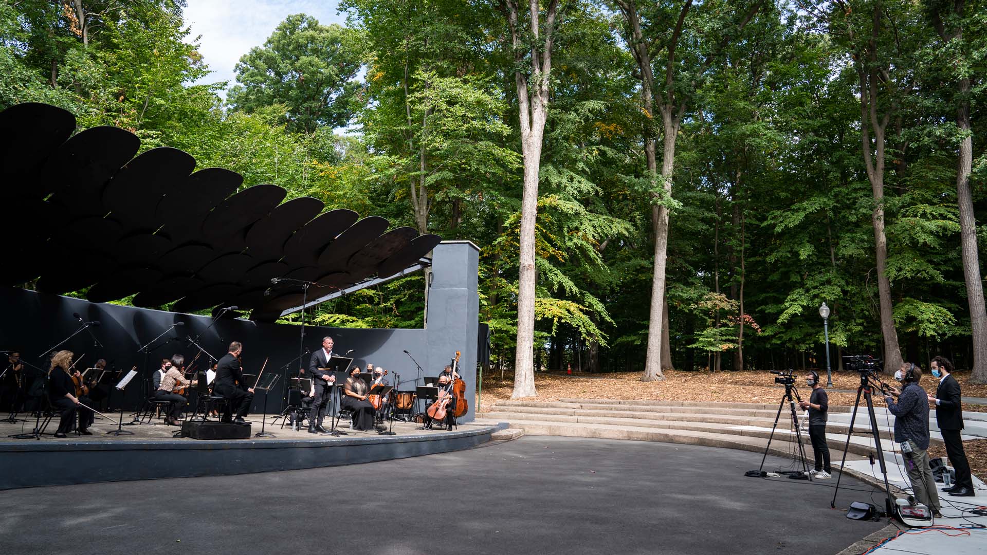 At left, Orpheus performs in a black bandshell in a sunlit, leafy green park while, at right, a masked and physically distanced film crew records the concert.