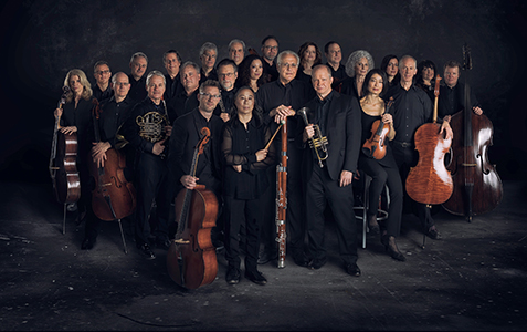 Orpheus Chamber Orchestra posed, standing with their instruments, in black clothing against a dark background